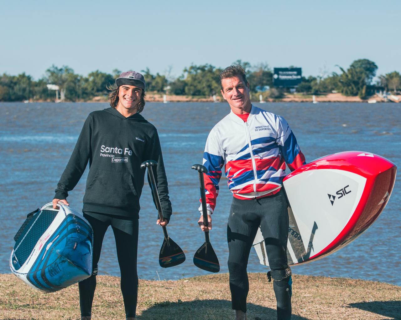 Santino junto a su tío y entrenador, Guivi Basaldella, parados con sus tablas y remos a orillas del agua